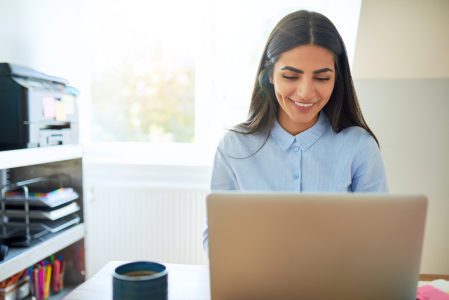 Single laughing young Indian woman beside printer on shelf and wearing long hair while seated in front of laptop computer in bright room